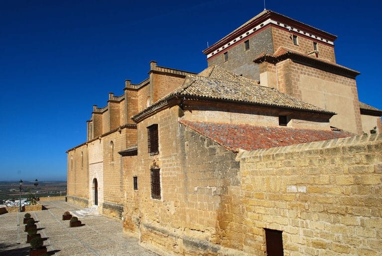Colegiata de Osuna vista lateral con muros de sillar dorado y torre cuadrada bajo un cielo azul intenso.