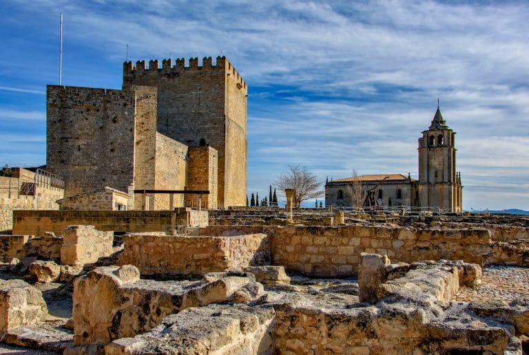 Fortaleza de la Mota en Alcalá la Real con antiguos muros y ruinas de piedra en primer plano y la iglesia renacentista al fondo bajo un cielo azul.