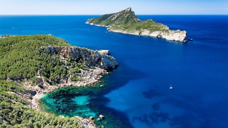 vista aérea de la isla dragonera frente a la costa de mallorca con el mar de un azul profundo y un velero solitario navegando.