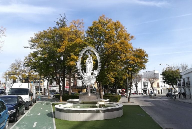 Monumento de la Virgen María Auxiliadora de metal blanco rodeado de árboles con hojas amarillas en una plaza urbana de Utrera.