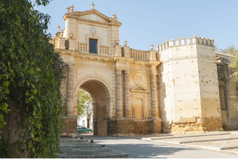 Puerta de Córdoba en la muralla de Carmona, un arco monumental de piedra con columnas y detalles ornamentales, flanqueado por torres almenadas y vegetación a los costados.