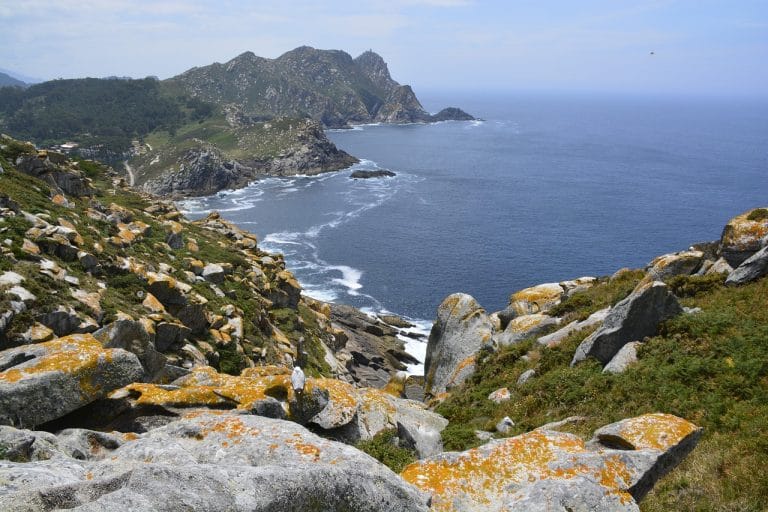 Impresionante paisaje costero de las Islas Cíes, Galicia, con acantilados rocosos, líquenes naranjas y el mar azul cristalino.