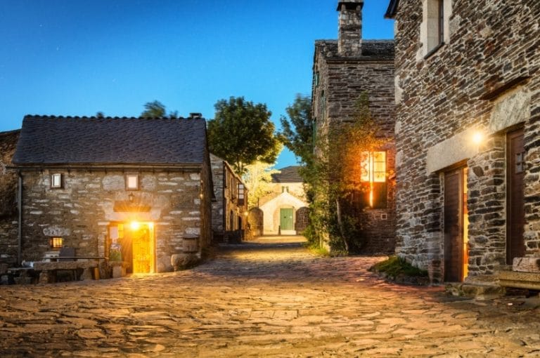 Calle empedrada al anochecer en O Cebreiro, Galicia, flanqueada por casas de piedra con luz cálida en las ventanas y faroles encendidos.