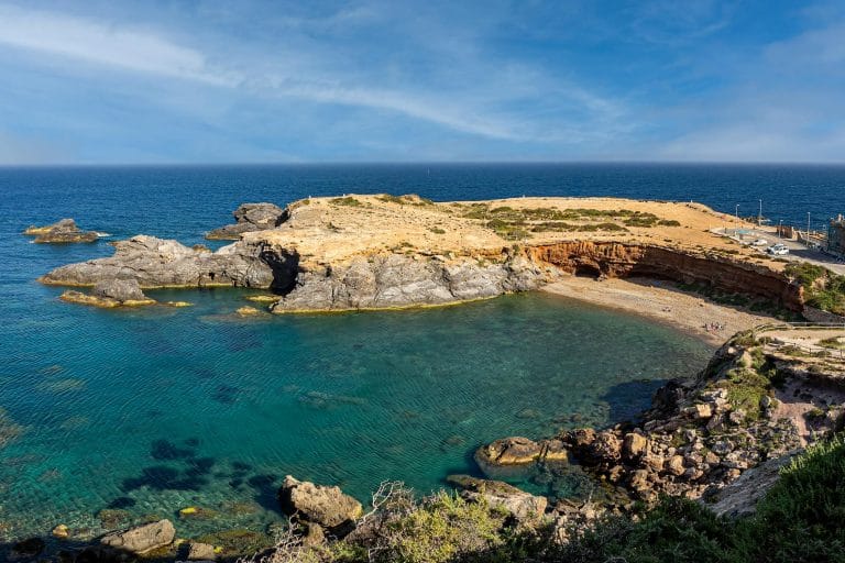 Vista panorámica de la Cala Cortina en Cartagena, con aguas cristalinas de color turquesa, formaciones rocosas y pequeña playa en un ensenada junto al mar.