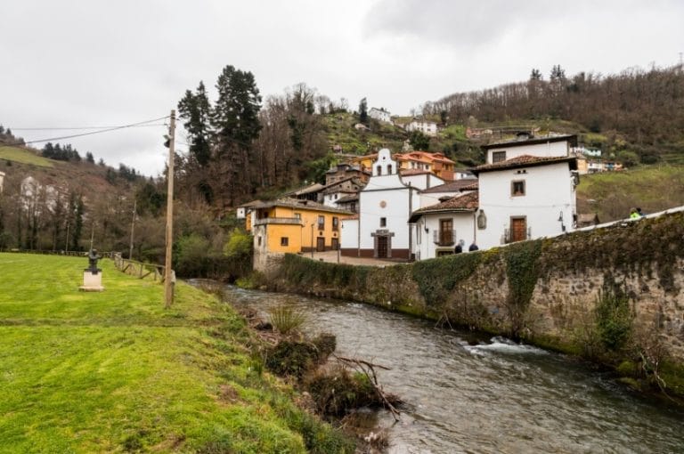 Casas tradicionales junto al río en una zona rural de Cangas del Narcea, con colinas verdes y paisaje de montaña al fondo.