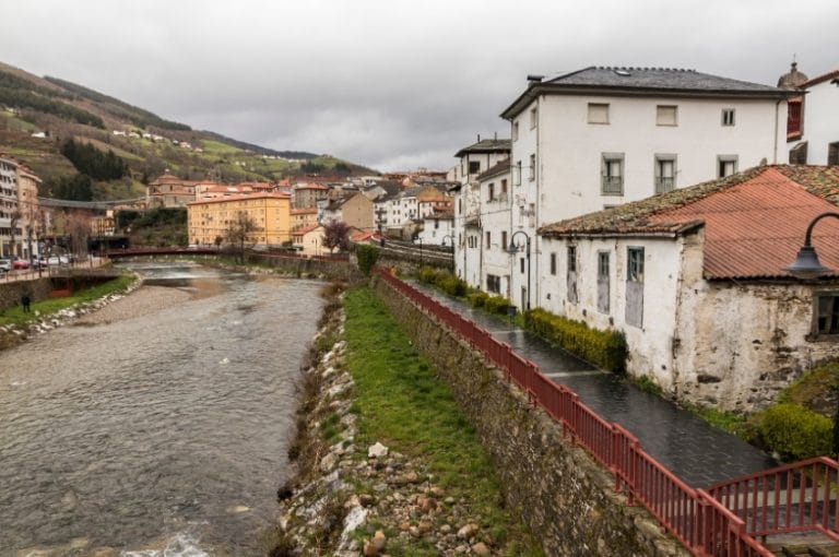 Vista del río Narcea a su paso por el casco urbano de Cangas del Narcea, con casas tradicionales y paisaje montañoso al fondo en un día nublado.