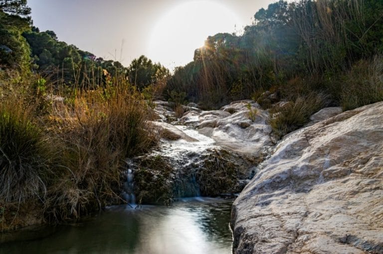 Paisaje natural en la fuente Caputa con agua que fluye entre rocas, vegetación y el sol al fondo iluminando el entorno
