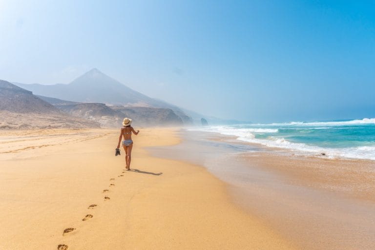 Mujer caminando descalza por la playa virgen de Cofete en Fuerteventura con montañas al fondo y aguas cristalinas del Atlántico.