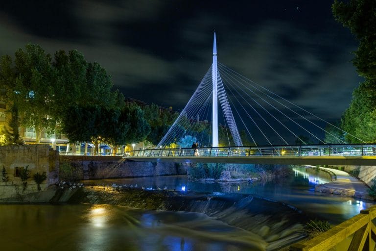 Pasarela moderna iluminada por la noche sobre el río Segura en Murcia, con un mástil central blanco y cables tensores reflejándose en el agua.