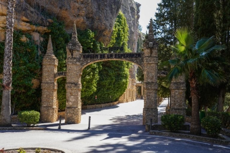 Entrada principal del santuario de nuestra señora de la esperanza en calasparra con un arco de piedra rodeado de palmeras y cipreses.