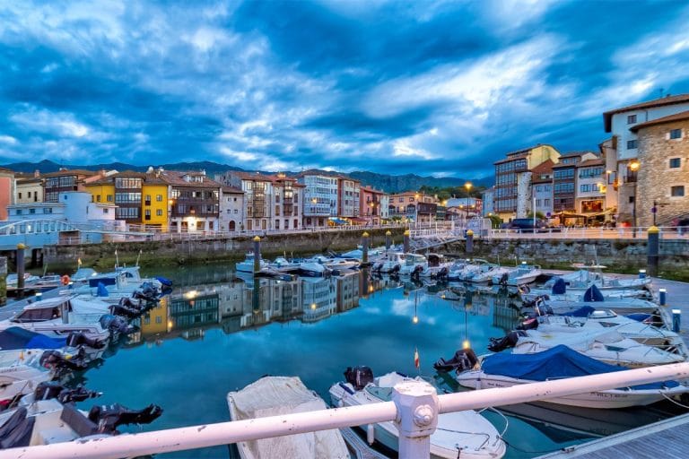 Vista del puerto de Llanes al atardecer con pequeñas embarcaciones amarradas y coloridas fachadas tradicionales reflejadas en el agua, bajo un cielo azul dramático en Asturias.