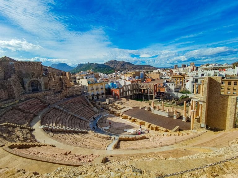 Vista aérea del Teatro Romano de Cartagena (Murcia) con sus gradas semicirculares excavadas en la roca, el escenario con columnas clásicas y, al fondo, el casco urbano de la ciudad y las colinas circundantes.
