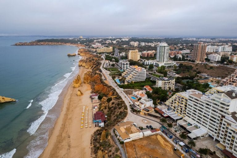 Vista aérea de la playa de Portimão con formaciones rocosas, sombrillas alineadas y edificios modernos junto al mar en el Algarve.
