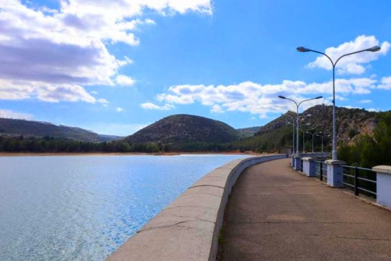 Paseo a lo largo del embalse de Gallipuén, con un muro de contención de hormigón y farolas que bordean el camino pavimentado. Las aguas tranquilas del embalse se extienden hacia colinas verdes bajo un cielo azul con nubes.