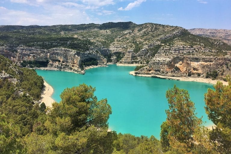 Vista panorámica del embalse de Santolea, mostrando sus aguas turquesas rodeadas de escarpados acantilados rocosos y colinas con vegetación, bajo un cielo azul despejado.