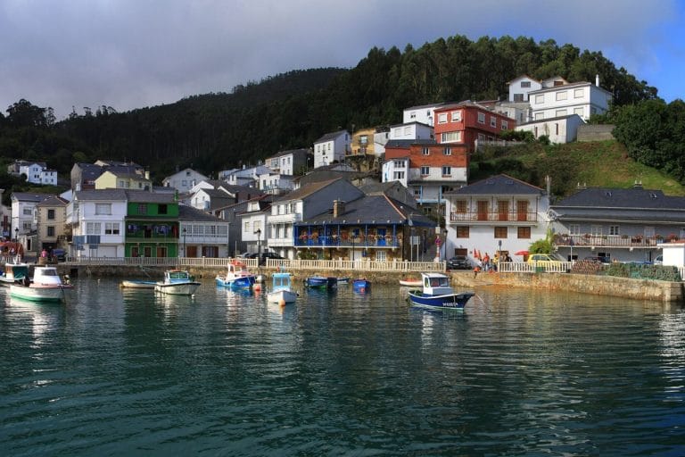 Vista panorámica del pintoresco puerto de O Barqueiro, Mañón, A Coruña, con casas de colores ascendiendo por la ladera de la montaña y pequeñas embarcaciones en la ría.