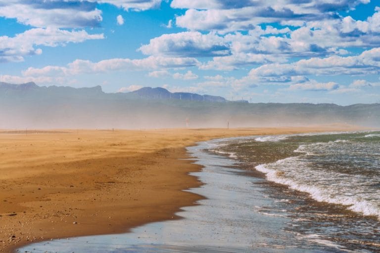 Playa del Fangar en el Delta del Ebro con olas suaves, arena dorada y vistas al faro entre la niebla