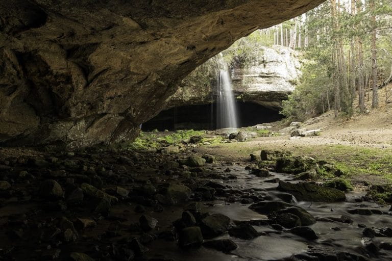 Vista de la Cascada de Duruelo, un pequeño salto de agua que cae sobre una gruta rocosa y un riachuelo, rodeado de un bosque de pinos