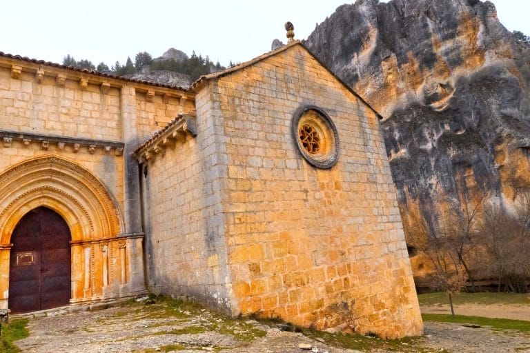 Vista exterior de la Ermita de San Bartolomé, un edificio románico de piedra con un arco en la puerta y un rosetón circular, construido al pie de un imponente acantilado rocoso